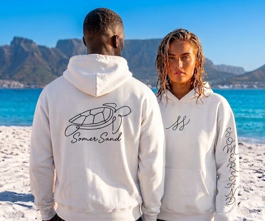 Two models wearing white SomrSand hoodies, one showing the turtle graphic on the back and the other showing the front chest logo, standing on a sandy beach with a scenic mountain and ocean background.