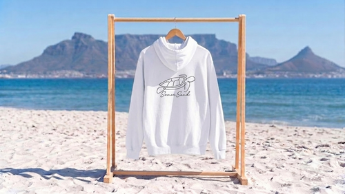 A white hooded sweatshirt with a black graphic and the words somersand hanging on a wooden frame on a sandy beach with the ocean and mountains in the background.