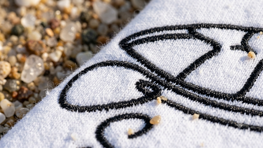 A close-up of a white textured clothing with a black embroidered cartoon-style outline, lying on small pebbles and sand.